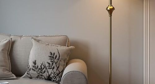 A classic antique brass standing light lamp with a cream shade, placed next to a cozy sofa in a living room.