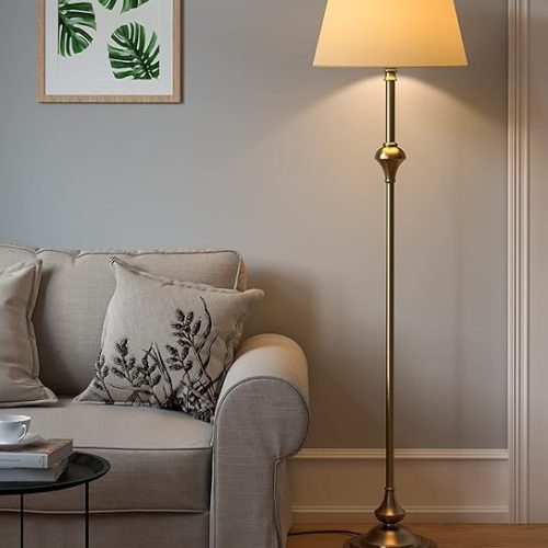 A classic antique brass standing light lamp with a cream shade, placed next to a cozy sofa in a living room.
